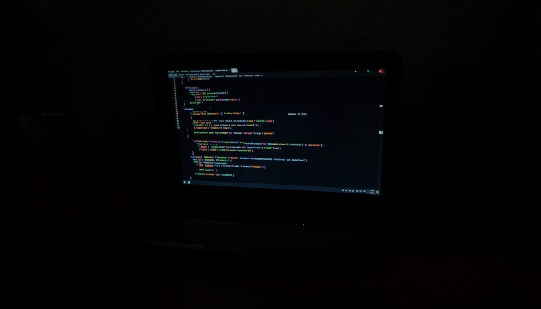 Dark low-key photograph of a laptop screen glowing with lines of code in a dim room, deep shadows across the desk